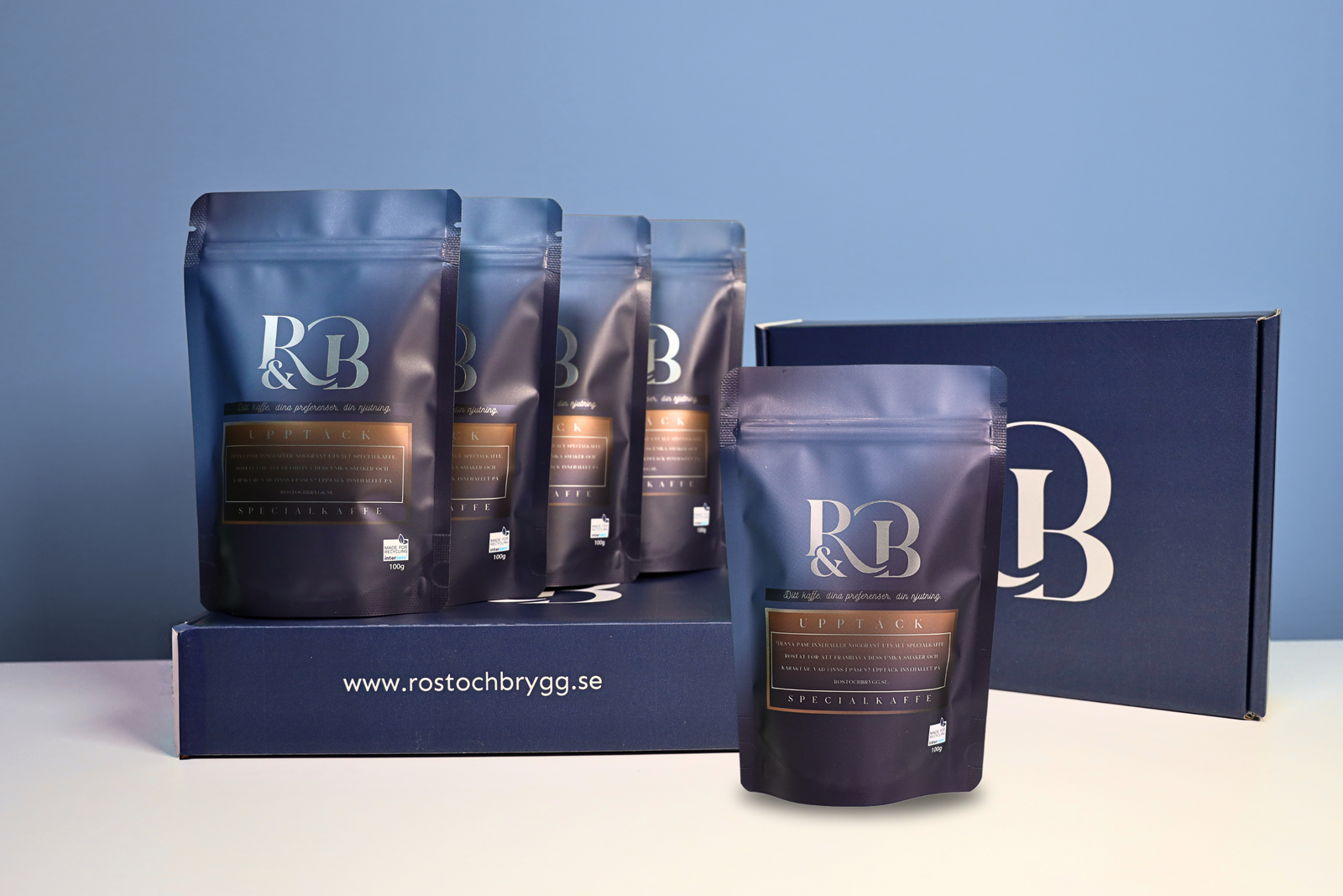 Fem påsar med specialkaffe från Rost & Brygg, uppställda framför en blå förpackningslåda med företagets logotyp och webbadress www.rostochbrygg.se. Paketet som visas kallas för Utforska.